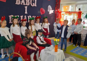 Hubert prezentuje dalszą część inscenizacji. Chłopiec stoi odwrócony w kierunku kanapy, na której siedzi Babcia i Dziadek i podnosi do góry bajkowe buty.