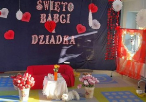 Dekoracja z okazji Dnia Babci i Dziadka - na granatowym tle przypięty jest napis "Święto Babci i Dziadka" oraz serduszka. Z prawej strony stoi fotobudka, a na szafce wazon z białymi gałązkami, na których wiszą serca. Na środku dywanu stoją kosze z balonowymi różami, kanapa przykryta czerwonym materiałem, stolik, na którym znajduje się wazon z tulipanami, filiżanki. Obok stolika siedzi pluszowy kotek.