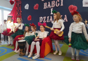 Dziewczynki ubrane w białe bluzeczki, zielone i czerwone spódniczki oraz opaski na głowach grają na instrumentach: gitarach, trójkącie, flecie, tamburynie, bębenku, trójkącie, grzechotkach. Trzy dziewczynki siedzą na kanapie, a cztery stoją obok kanapy.