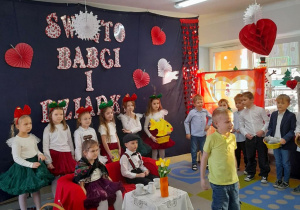 Na środku sceny stoi Fabian. Chłopiec mówi fragment inscenizacji "Prezenty dla Babci i Dziadka". W tle dekoracja oraz koledzy i koleżanki z grupy.