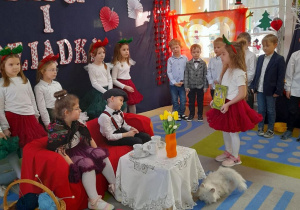 Babcia i Dziadek siedzą na kanapie, a przed nimi stoi stolik nakryty białą serwetką, na którym znajdują się filiżanki i wazon z kwiatkami. Amanda stoi przed Dziadkiem i wręcza mu książkę z bajkami.