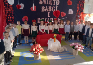 Dzieci stoją w półkolu na tle dekoracji. Na środku dywanu stoją kosze z balonowymi różami, kanapa przykryta czerwonym materiałem, stolik, na którym znajduje się wazon z tulipanami, filiżanki. Obok stolika siedzi pluszowy kotek.