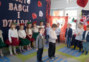 Bartek i Marcel stoją na środku sceny i recytują fragment wiersza dla Babci i Dziadka. W tle koledzy i koleżanki stoją w półkolu oraz dekoracja.