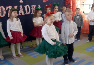 Wiktor i Zuzia recytują fragment wiersza pt. "Życzenia". W tle grupka dzieci oraz dekoracja.