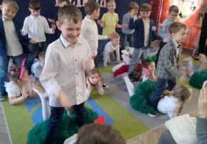 Chłopcy stoją, a dziewczynki leżą na dywanie poruszając nogami podczas tańca.