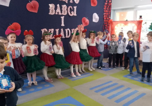 Dzieci stoją na dywanie w półkolu i trzymają przed sobą upominki - portrety. W tle dekoracja.