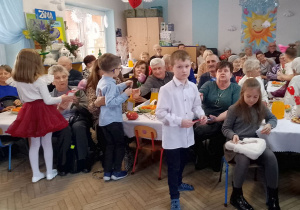 Babcie i Dziadkowie siedzą przy udekorowanych stołach, na których znajduje się poczęstunek, a dzieci wręczają swoim bliskim upominki.