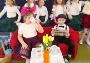 Nadia z Mikołajem siedzą na kanapie. Dziewczynka w roli Babci pije herbatę z filiżanki, a chłopiec w roli Dziadka czyta gazetę. Za kanapą stoją dziewczynki z grupy.