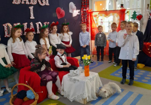 Antoś stoi obok kanapy, na której siedzi Babcia i Dziadek. Chłopiec trzyma w ręku stoliczek bajkowy dla Babci i odtwarza fragment inscenizacji. W tle grupka rówieśników oraz dekoracja.