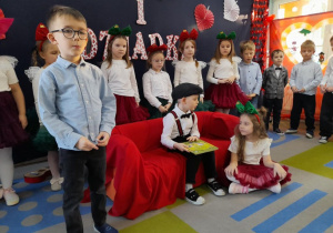 Mikołaj stoi obok kanapy w roli narratora. Na kanapie siedzi Dziadek i opowiada wnuczce bajkę. W tle grupka rówieśników stoi obok siebie.