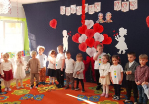 Grupa "Motylków" ustawiona w półkolu na tle dekoracji, na której znajduje się bukiet z serduszek, chłopiec z dziewczynką i napis kochamy was, za nimi pani Grażynka, która wita zaproszonych gości.