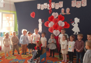 Dzieci ustawione w półkolu śpiewają piosenkę , w środku w fotelu siedzi dziewczynka przebrana za babcię i robi na drutach.