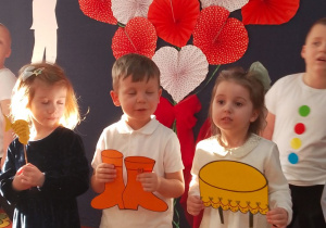 Dwie dziewczynki i chłopiec trzymając w rękach rekwizyty mówią wiersz.