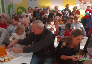 Chłopiec wręcza swoim babciom upominki, pozostałe dzieci ze swoimi babciami i dziadkami w trakcie poczęstunku.