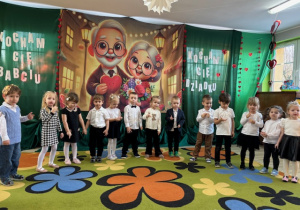 "Pszczółki" stoją w półokręgu na tle dekoracji z okazji Dnia Babci i Dziadka.