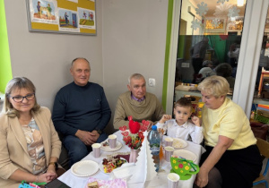 Przy stoliczku z poczęstunkiem siedzi Olaf i jego babcie oraz dziadkowie.