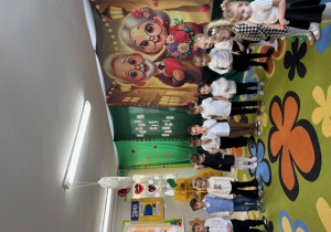 Dzieci stoją w półokręgu na tle dekoracji z okazji Dnia Babci i Dziadka.