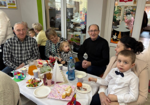 Przy stoliczku z poczęstunkiem siedzi Michałek z babcią oraz Alicja z dziadkami.