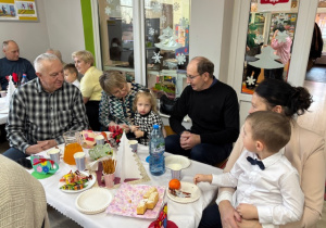Przy stoliczku z poczęstunkiem siedzi Michałek z babcią oraz Alicja z dziadkami.