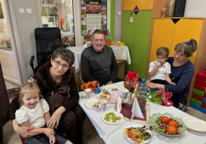 Przy stoliczku z poczęstunkiem siedzi Maja i Nikodem wraz z babciami i dziadkiem.