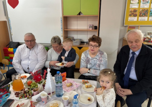 Przy stoliczku z poczęstunkiem siedzi Marysia i Oluś ze swoimi dziadkami.