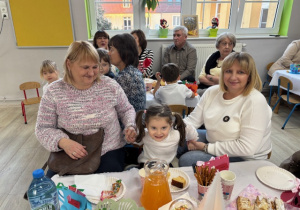 Przy stoliczku z poczęstunkiem siedzi Hania, a po jej dwóch stronach babcie.