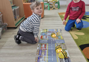 Olek i Julek siedzą przed matą do kodowania z wizerunkiem drogi i obserwują jadące po niej BeeBoty.