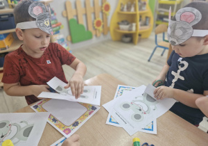 Franek i Szymon siedząc przy stoliku wycinają z papieru wizerunki myszy.