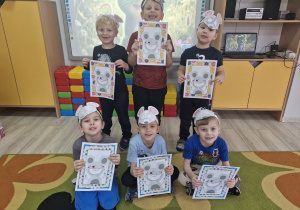 Dzieci stoją przed tablicą multimedialną. Chłopcy maja na głowach opaski z wizerunkami myszy, a w dłoniach trzymają wykonane przez siebie prace z szarą myszką.