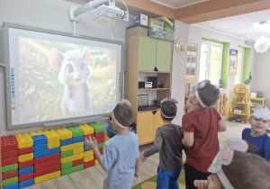 Dzieci stoją przed tablicą multimedialną do piosenki o szarej myszce.