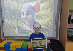 Olek stoi przed tablicą multimedialną, na której wyświetlony jest kard z piosenki "Szara myszka". Chłopiec trzyma w dłoniach pasujący do wyświetlanego zdjęcia obrazek.