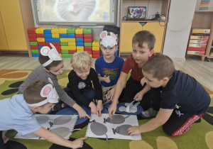 Dzieci siedzą na dywanie i przeliczają papierowe myszki leżące na białych kartonach.