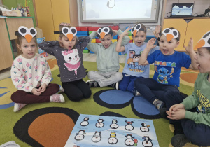 12. Dzieci siedzą na dywanie, przed nimi leży rozłożony plakat z pingwinami. Przedszkolaki mają otwarte usta i dłonie uniesione w górę.