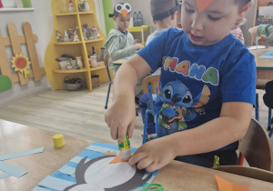 8. Szymek siedzi przy stoliku. Chłopiec przykleja wycięte fragmenty pingwinka do niebieskiego kartonu.