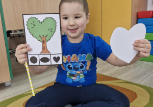 8. Szymek trzyma w dłoni obrazek z wizerunkiem drzewa w kształcie serduszka, a w drugiej dłoni serduszko.