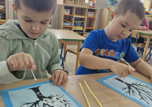 9. Stanisław i Szymon siedzą przy stoliku i malują farbą plakatową wizerunki drzew na niebieskim papierze.