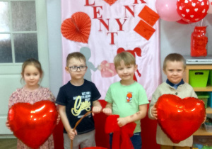 Czworo dzieci pozuje do zdjęcia na tle dekoracji walentynkowej. Franek i Józio trzymają koszyk, w którym znajdują się pudełeczka walentynkowe z niespodzianką. Po bokach stoją Filip i Felicja z balonami w rękach.