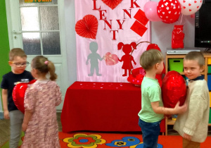 Dzieci tańczą w parach trzymając balon w kształcie serca między brzuchami. W tle dekoracja walentynkowa.