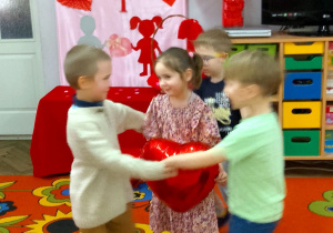 Chłopcy utworzyli małe kółeczko, a w środku tańczy Felicja z balonem w kształcie serca. W tle dekoracja walentynkowa.