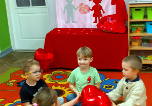 Czworo dzieci siedzi na dywanie w małym kółeczku i podaje sobie balon w kształcie serca przy muzyce. W tle dekoracja: na różowym materiale przypięty jest napis Walentynki oraz sylwety dzieci, serduszek, kopert. Na szafce przykrytej czerwonym materiałem stoi koszyk w walentynkowymi upominkami. Obok na szafce stoi wazon z balonami w serduszka.