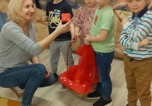 Filip, Józio, Felicja i Franek stoją z koszykiem, w którym znajdują się walentynkowe pudełeczka z niespodzianką. Przedszkolaki wręczyły upominek Pani Agnieszce, która przykucnęła obok dzieci.
