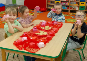 Dzieci z grupy „Motylków” siedzą przy udekorowanym stole i trzymają swoje upominki. Przed nimi leżą walentynkowe pudełeczka oraz ciastka w kształcie serca na talerzykach.
