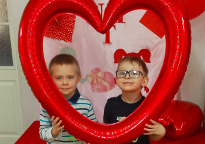 Filip i Franek stoją obok siebie na tle dekoracji. Chłopcy trzymają balon w kształcie serca.