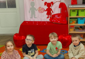 Dzieci siedzą na dywanie, a przed nimi leżą sylwety babeczek z kartonu i serduszka. W tle dekoracja walentynkowa.