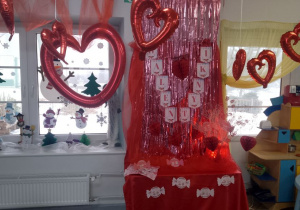Dekoracja z okazji Walentynek: na czerwonym materiale przypięty napis Walentynki oraz czerwone serca. Na szafce pokrytej czerwonym materiałem stoi wazon z białymi gałęziami i czerwonymi serduszkami oraz koszyczek z lizakami. Nad dekoracją wiszą czerwone balony w kształcie serc