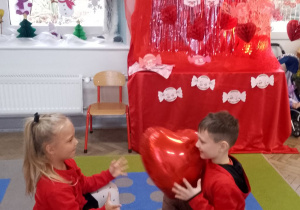 Tosia i Antek siedzą na dywanie i podają sobie balonowe czerwone serce.