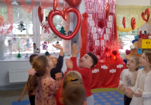 Dzieci stoją w rozsypce i podrzucają balonowe serce. W tle dekoracja walentynkowa.