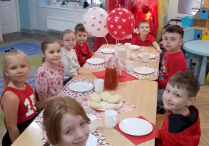 Dzieci siedzą przy wspólnym stole i spożywają posiłek. Na stolikach znajduje się wazonik z czerwonymi balonami w serduszka oraz serwetki w serduszka.
