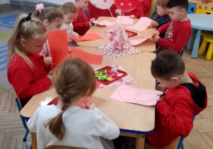 Dzieci siedzą przy wspólnym stole i wykonują kartki walentynkowe w kształcie serc. Na stole znajduje się wazonik z czerwonymi balonami w serduszka, papierowe serca, klej oraz ozdoby walentynkowe.