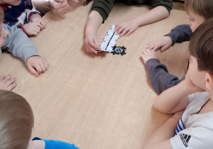 Grupka dzieci siedzi przy stoliku i układają z elementów trasę do Ozobotów. Obok kuca nauczycielka ze Szkoły Podstawowej nr 6 trzymająca telefon komórkowy.